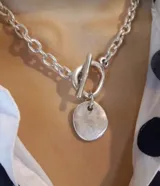 Collar de eslabones gruesos plateados con cierre tipo toggle (barra y argolla) y un dije colgante de placa metálica irregular.