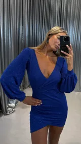 Vestido corto de fiesta en color azul eléctrico con acabado brillante. Presenta escote en V cruzado, mangas largas abullonadas con puños ajustados y diseño drapeado en la cintura que crea un efecto envolvente en la falda.