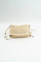 Cartera bandolera de tejido artesanal en color beige, con solapa frontal y correa larga ajustable decorada con cuentas de madera. Incluye detalles de flecos laterales con cuentas.
