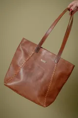 Bolso tipo shopper de cuero color suela con textura grabada, detalle de costuras laterales trenzadas y placa metálica con logo de la marca. Cuenta con doble asa larga para colgar al hombro, cierre principal y múltiples bolsillos internos y externos.