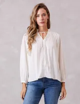 Blusa de gasa con plisado soleil, cuello a la base fruncido con escote en V regulable mediante lazos, y mangas largas abullonadas con puños sastreros.