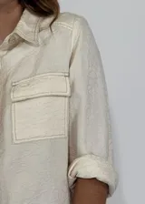 Chaqueta color crudo con cuello camisero, cierre frontal con botones, mangas 3/4 con puños arremangados y dos bolsillos de parche en el pecho.