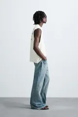 Camiseta sin mangas (manga sisa) de punto color crema, corte relaxed fit, con cuello redondo y acabados irregulares en las sisas. Presenta un texto estampado en el pecho que dice "SERIAL N. 8763/ 7.3Z".