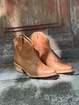 Botas de cuero color suela, de estilo texano, con caña baja, puntera fina y detalles de costuras decorativas.