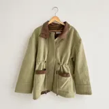 Chaqueta de silueta relajada, confeccionada en paño de lana color verde militar con detalles en pana de algodón marrón en el cuello y los bolsillos. Cuenta con cuello alto, bolsillos frontales amplios y cordón regulable en la cintura.