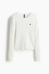 Buzo de punto trenzado color blanco, con cuello redondo, manga larga y un pequeño corazón bordado en el pecho. Presenta terminaciones acanaladas en cuello, puños y ruedo.