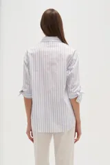 Camisa de algodón con rayas verticales azules y blancas, corte ancho y manga larga con lazos ajustables en los puños. Presenta un bordado de dos palmeras en color rosa brillante en el área del pecho.