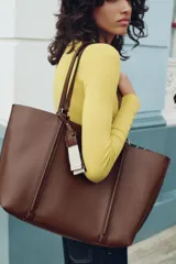 Bolso tipo shopper de color marrón, con diseño minimalista, doble asa de hombro y cierre magnético. Incluye un colgante decorativo con detalle de cinta en tonos claros y un bolsillo interior amplio.