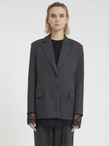 Blazer de mujer color gris oscuro con un sutil patrón de cuadros. Presenta solapas clásicas, cierre frontal de dos botones y bolsillos con tapa. Se combina con un top negro de encaje y pantalones a juego.