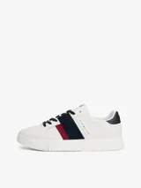Zapatillas urbanas Tommy Hilfiger de corte bajo, confeccionadas en piel lisa color blanco. Presentan paneles laterales en azul marino y rojo, cordones oscuros y suela de goma gruesa con diseño dentado. Incluyen detalles de la marca en la lengüeta, lateral y talón.