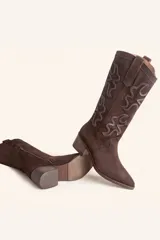 Bota de caña alta estilo tejano confeccionada en gamuza color marrón oscuro. Presenta detalles de costuras decorativas en forma de llama en la caña, taco cuadrado de madera y punta almendrada.