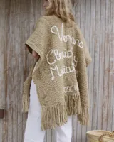 Ruana de lana merino tejida a mano, color beige, con diseño amplio y flecos en el ruedo. Presenta un bordado en hilo blanco en la espalda con la frase 'Verano, clericot y medialunas'.