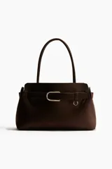Bolso tote de acabado suave, color marrón oscuro, con asas tubulares y detalle de correa frontal con hebilla metálica dorada.