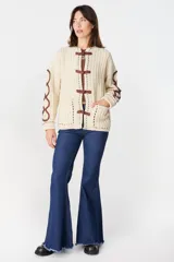 Cardigan de punto color crudo con textura acanalada, cierre frontal mediante alamares de color marrón y detalles decorativos de cordones en zigzag en las mangas. Presenta bolsillos frontales y costuras con pespuntes en contraste.