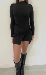 Vestido corto negro de manga larga, con cuello alto y diseño ajustado. Presenta un detalle de nudo drapeado en la parte frontal de la falda, creando un efecto de tajo lateral.