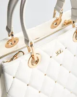 Cartera tipo bolso de mano XL color blanco de símil cuero con diseño matelassé romboidal, tachas metálicas doradas, doble asa corta y correa bandolera regulable y desmontable. Incluye herrajes dorados y logo metálico de la marca en el frente.
