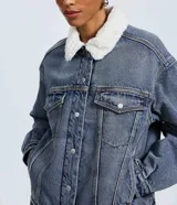 Campera trucker de jean azul con cuello de corderito blanco, cierre frontal con botones metálicos, bolsillos frontales con solapa y puños abotonados.
