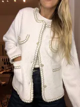 Saco tipo chaqueta de punto texturizado en color blanco, con diseño inspirado en el estilo tweed. Presenta bordes decorados con una trenza metálica dorada y plateada, cierre frontal con botones perlados, bolsillos frontales y detalles de solapas falsas en el pecho.