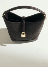 Bolso tipo bucket de tamaño pequeño, confeccionado con tejido de rafia color marrón con efecto texturizado y detalles en cuero sintético liso del mismo tono. Cuenta con un asa corta superior, herrajes metálicos dorados, cierre de imán y un detalle decorativo con candado dorado en la parte frontal.