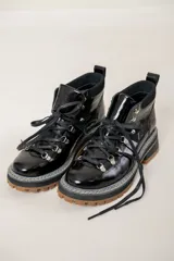 Borcegos de cuero negro con acabado charolado y textura efecto cocodrilo en los laterales. Presentan un diseño acordonado con ganchos metálicos, suela track de goma en color caramelo y costuras reforzadas en el borde.