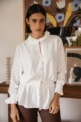 Blusa blanca de manga larga con cuello redondo estilo bebé, cierre frontal con botones y detalle de volados en puños y cintura.