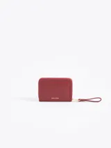 Billetera compacta de cuero vacuno color bordó, con cierre perimetral, correa de muñeca y aplique metálico dorado en forma de mariquita en el frente.