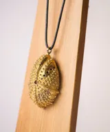 Collar con dije en forma de erizo de mar con textura granulada, suspendido de un cordón negro. El dije presenta un diseño orgánico con pequeños orificios y una superficie detallada.