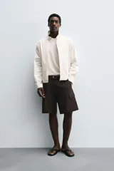 Bermuda de corte regular fit color marrón oscuro, confeccionada en tejido de lyocell y algodón. Presenta bolsillos cargo con solapa en las perneras, bolsillos delanteros y bolsillos traseros de vivo. Cierre frontal con cremallera y botón.