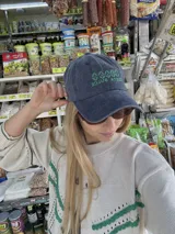 Gorro de visera de tela color gris desgastado, con bordado frontal en hilo rojo que dice "COSTA DULCE" y un diseño de ondas o símbolos repetidos encima.