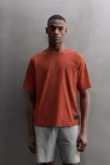 Camiseta de ajuste relajado (relaxed fit) color terracota, confeccionada en tejido técnico. Presenta cuello redondo, mangas ranglán cortas y un parche negro con logo en el bajo.