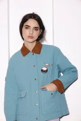 Campera celeste con cuello y puños de pana color marrón. Tiene cierre con botones a presión dorados, bolsillos frontales y parches bordados en el pecho.