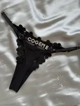 Tanga regulable negra de encaje con detalles de tiras elásticas, argollas metálicas y aplique de letras de strass con la frase 'COGEME'.