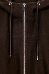 Chaqueta estilo bomber de corte oversize en color marrón oscuro, confeccionada en tejido suave. Presenta capucha con cordones ajustables, cierre frontal con cremallera, mangas largas con puños elásticos y bolsillos delanteros de vivo. El bajo cuenta con detalle de pliegues.