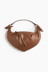 Bolso de hombro marrón con forma de media luna, confeccionado en cuero sintético. Presenta asa corta con nudos decorativos y cierre superior con tiras largas con tachuelas.