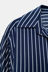 Camisa de rayas verticales azules y blancas, de cuello solapa con escote en V y cierre frontal con botones. Presenta mangas abullonadas por debajo del codo.