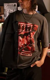 Remera de manga corta con cuello redondo y calce oversize, color gris oscuro. Presenta un estampado frontal con estética retro que incluye figuras humanas y una computadora antigua en tonos rojos.
