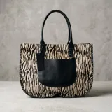 Cartera tipo tote con diseño de estampado animal print de cebra, combinada con un bolsillo frontal y asas en cuero negro. Presenta detalles de costuras artesanales en los bordes laterales y una etiqueta colgante decorativa.