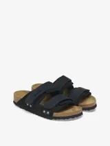 Sandalias Birkenstock modelo Uji de color negro, con dos tiras anchas ajustables mediante cierres de contacto y detalles de remaches metálicos. Confeccionadas con una combinación de gamuza y cuero nobuk, presentan la clásica plantilla anatómica de corcho y suela de goma negra.
