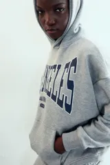 Sudadera gris con capucha y manga larga. Presenta un bolsillo canguro en la parte delantera y un estampado en azul marino con el texto "LOS ANGELES CALIFORNIA EST. 1975". Los acabados son de rib.