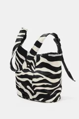 Bolso mini tipo saca asimétrica confeccionado en piel con pelo y estampado animal de cebra. Tiene asa de hombro ajustable, bolsillo interior con cremallera y cierre mediante imán.