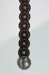Cinto ancho de cuero marrón con diseño de piezas circulares unidas, tachas doradas y una gran hebilla metálica circular con grabado ornamental.