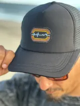 Gorra trucker negra con paneles frontales rígidos y paneles traseros de malla. Presenta un parche bordado en el frente con un diseño abstracto en tonos naranja, amarillo y blanco.