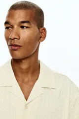 Camisa de manga corta con cuello abierto, confeccionada en tejido con textura. Presenta un corte holgado y bajo recto con aberturas laterales.