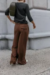 Pantalón de vestir marrón oscuro, tiro alto, con pernera ancha y pinzas frontales. Presenta un bolsillo con tapeta en la parte superior delantera.