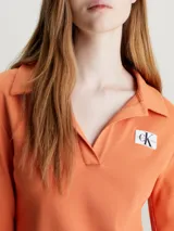Vestido corto naranja de punto de algodón elástico con textura de canalé. Tiene cuello polo en V, mangas largas acampanadas e insignia con el monograma de Calvin Klein en el pecho.