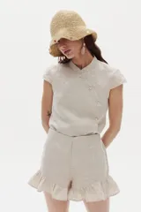 Camisa de lino color beige, con cuello mao y cierre cruzado al costado mediante botones de nácar. Presenta mangas cortas y diseño entallado.