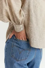 Blusa de manga larga con diseño de hombros abullonados y cuello redondo pequeño. Presenta cierre frontal con botones y un estampado fino de puntos sobre fondo beige.