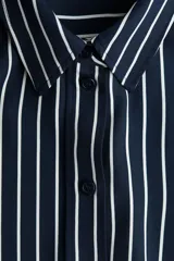 Camisa de manga larga con cuello clásico y cierre frontal de botones. Presenta un estampado de rayas verticales finas blancas sobre fondo azul marino. Los puños son anchos y también tienen rayas horizontales.