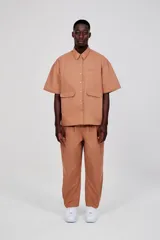 Sobrecamisa unisex color marrón claro, de manga corta y corte ancho. Presenta abertura frontal con botones tono sobre tono, dos bolsillos de parche en la parte inferior delantera y un bolsillo de ribete en el pecho. Logotipo de la marca estampado en contraste en el pecho y estampado grande en dos tonos en la espalda.