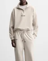 Conjunto de dos piezas compuesto por un buzo de cuello alto con cierre frontal corto y un pantalón tipo jogger de corte relajado. Ambas prendas presentan el logo RonaStudio bordado en color negro y están confeccionadas en algodón premium de tono beige.